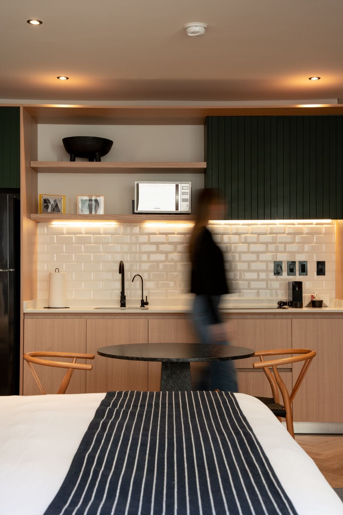 A suite kitchen with white tiles, warm wood and a guest walking past